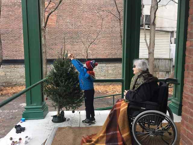 Photo of Sharon Holcroft sitting on the porch of the Fulford Residence with her grandson placing an item on a small spruce tree.